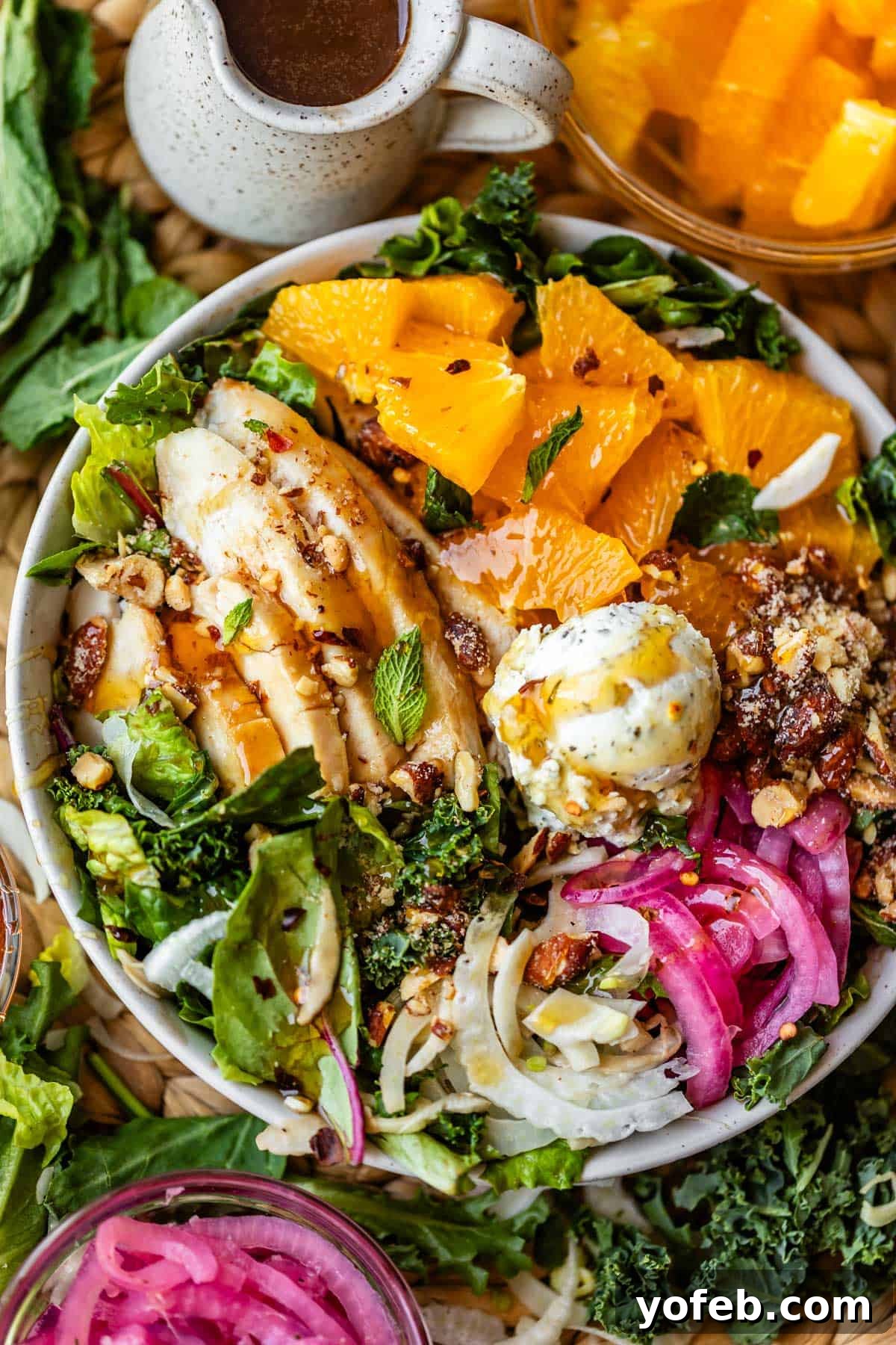 Mendocino Copycat Citrus and Goat Cheese Salad 2 A colorful salad bowl with sliced grilled chicken, orange segments, mixed greens, pickled onions, cheese, nuts, and fresh herbs, served with a small jug of dressing and a bowl of orange slices nearby.