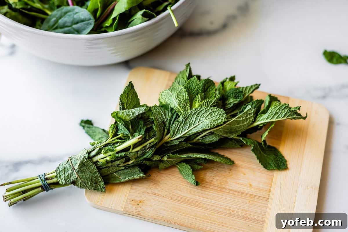 Mendocino Copycat Citrus and Goat Cheese Salad 11 A bunch of fresh mint leaves tied with a rubber band is placed on a wooden cutting board, with a bowl of leafy greens in the background on a marble countertop.