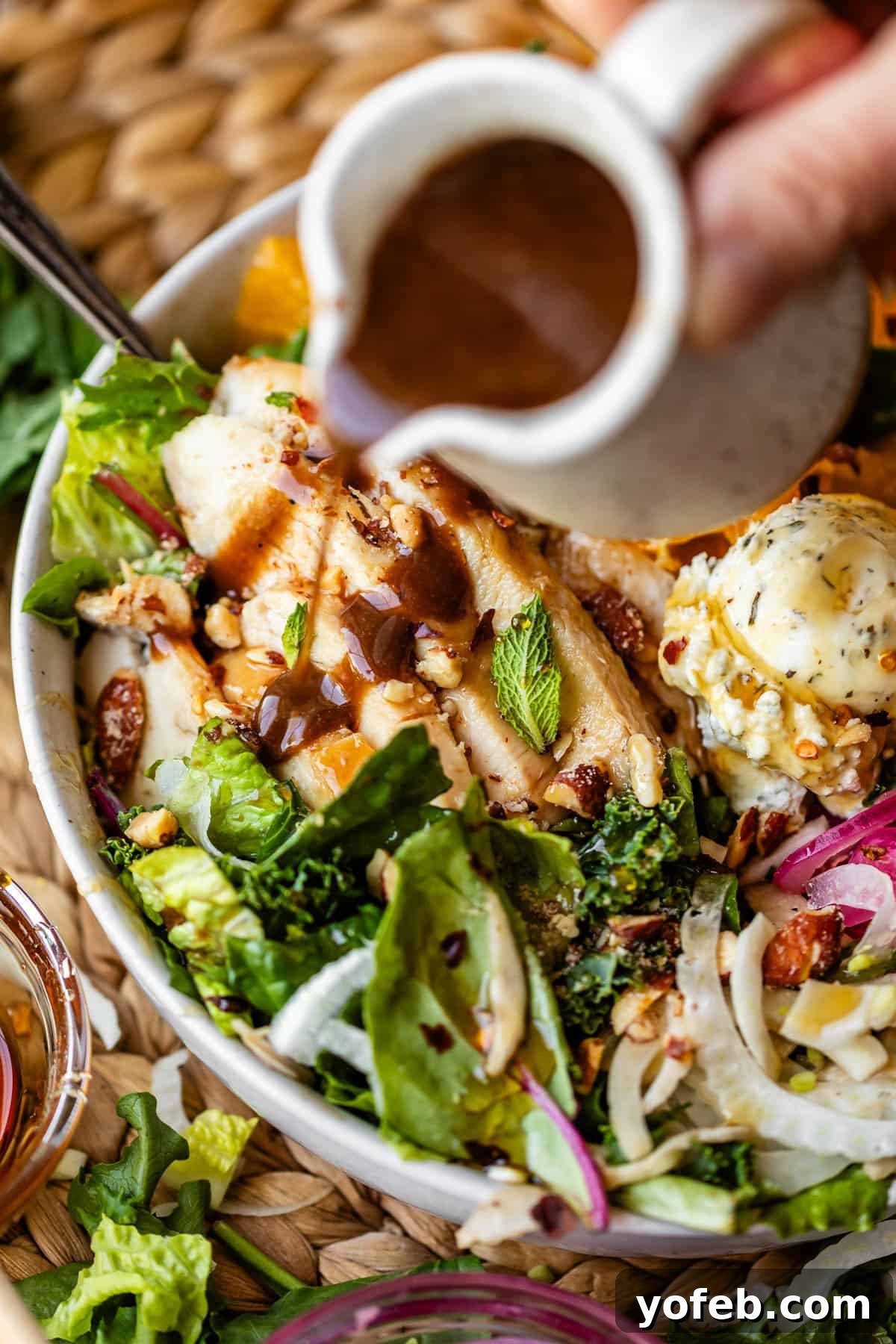 Mendocino Copycat Citrus and Goat Cheese Salad 4 A hand pours dark dressing over a salad bowl with grilled chicken, greens, red onions, orange slices, nuts, and a cheese-topped ball on a woven mat.