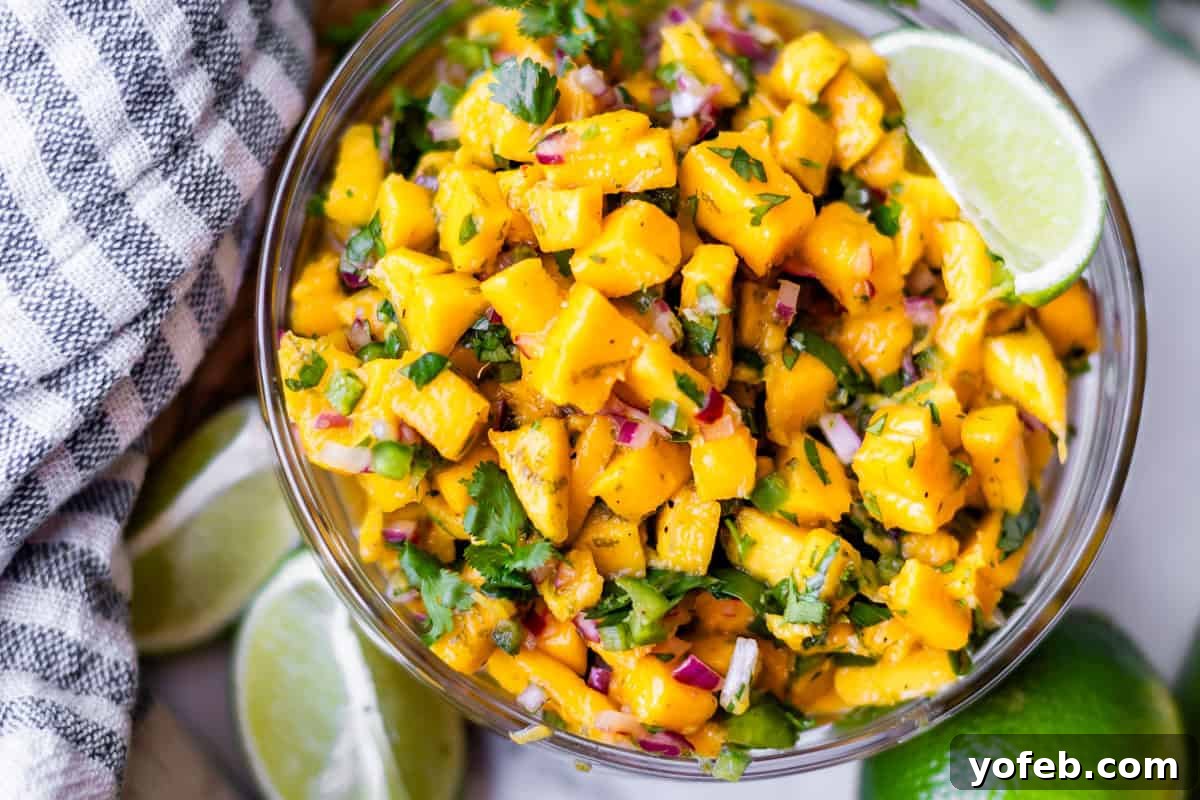 Zesty Mango Salsa Recipe with Lime and Cilantro 16 A glass bowl filled with vibrant mango salsa made of diced mango, red onion, cilantro, and lime wedges, sits next to a striped towel and more lime slices.