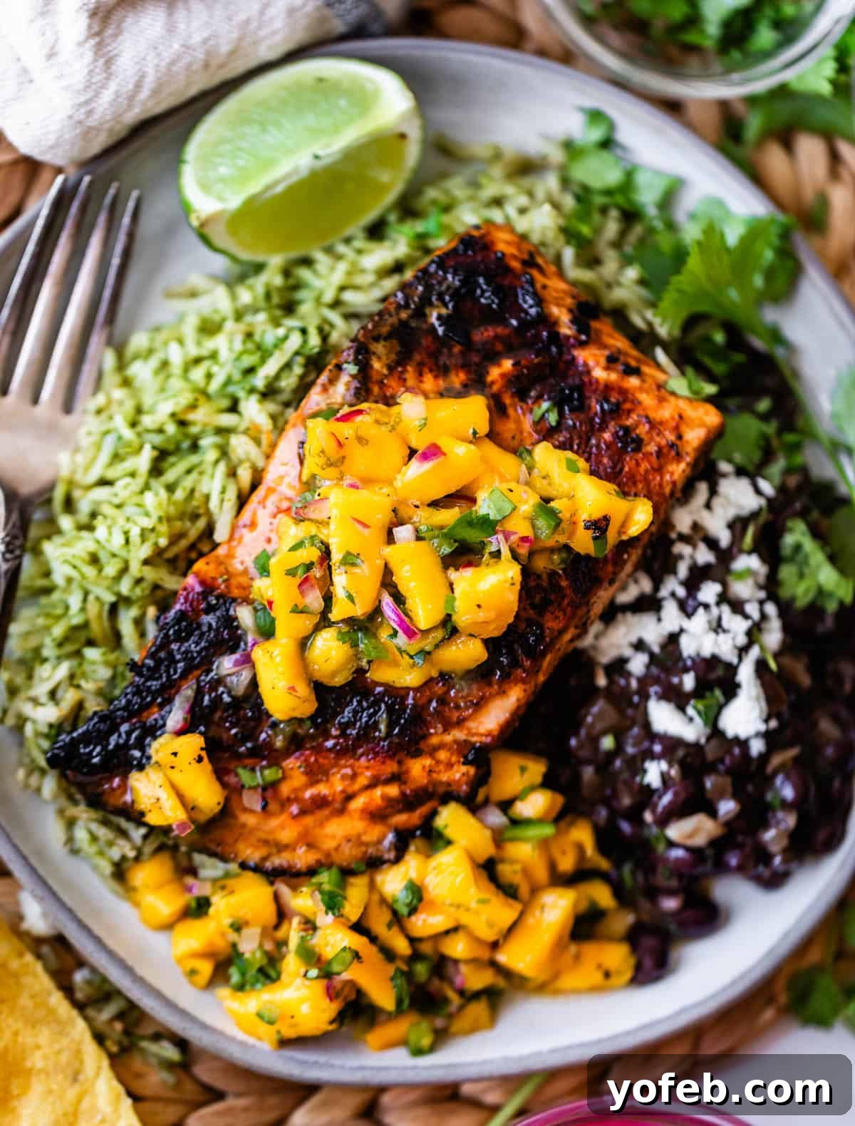 Zesty Mango Salsa Recipe with Lime and Cilantro 7 A plate with grilled salmon topped with mango salsa, served over green rice, with black beans, crumbled cheese, cilantro, and a lime wedge.