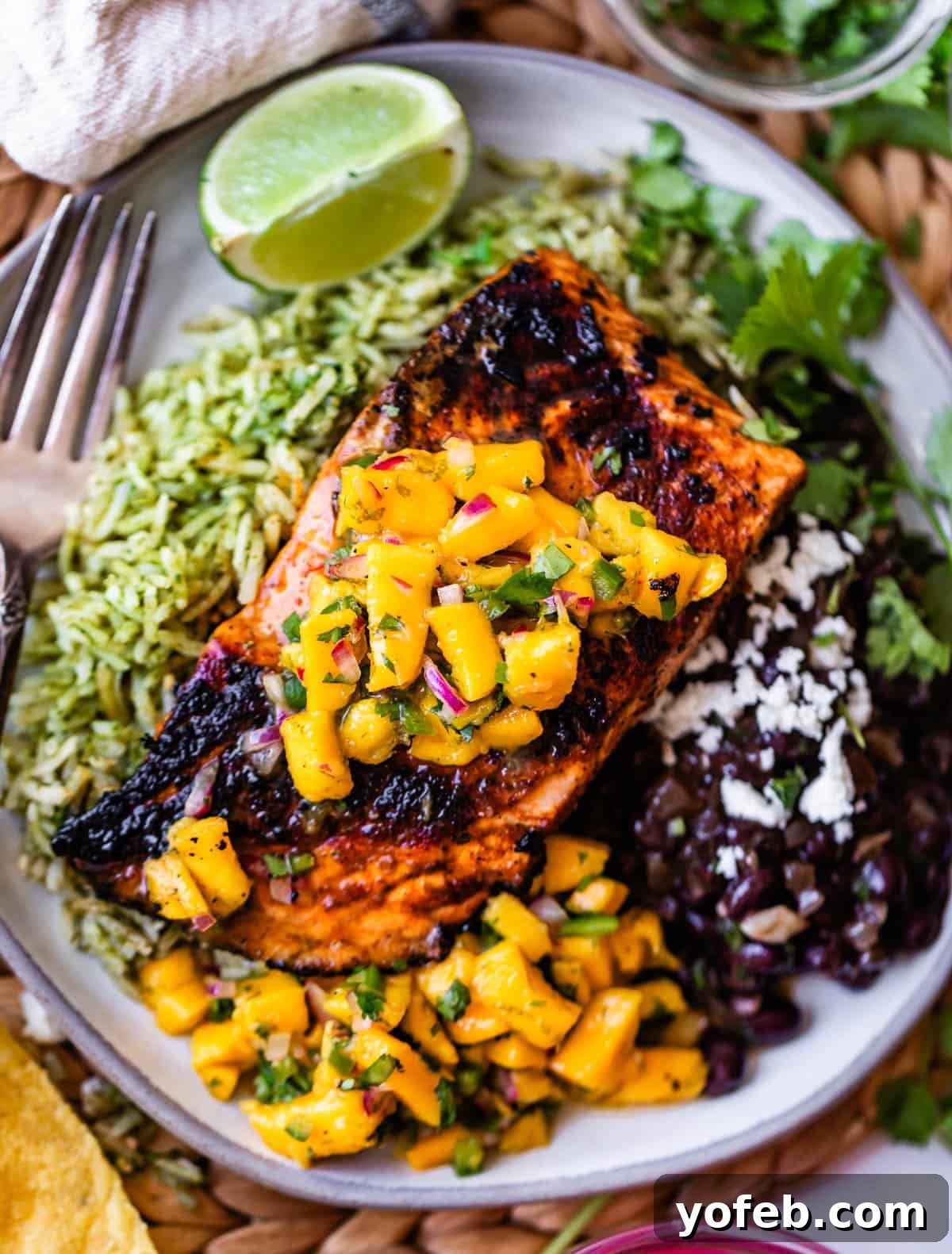 Chili-Lime Blackened Salmon 4 A plate of grilled salmon topped with mango salsa, served with green rice, black beans with cheese, a lime wedge, and fresh cilantro. A fork and tortilla are visible beside the plate.