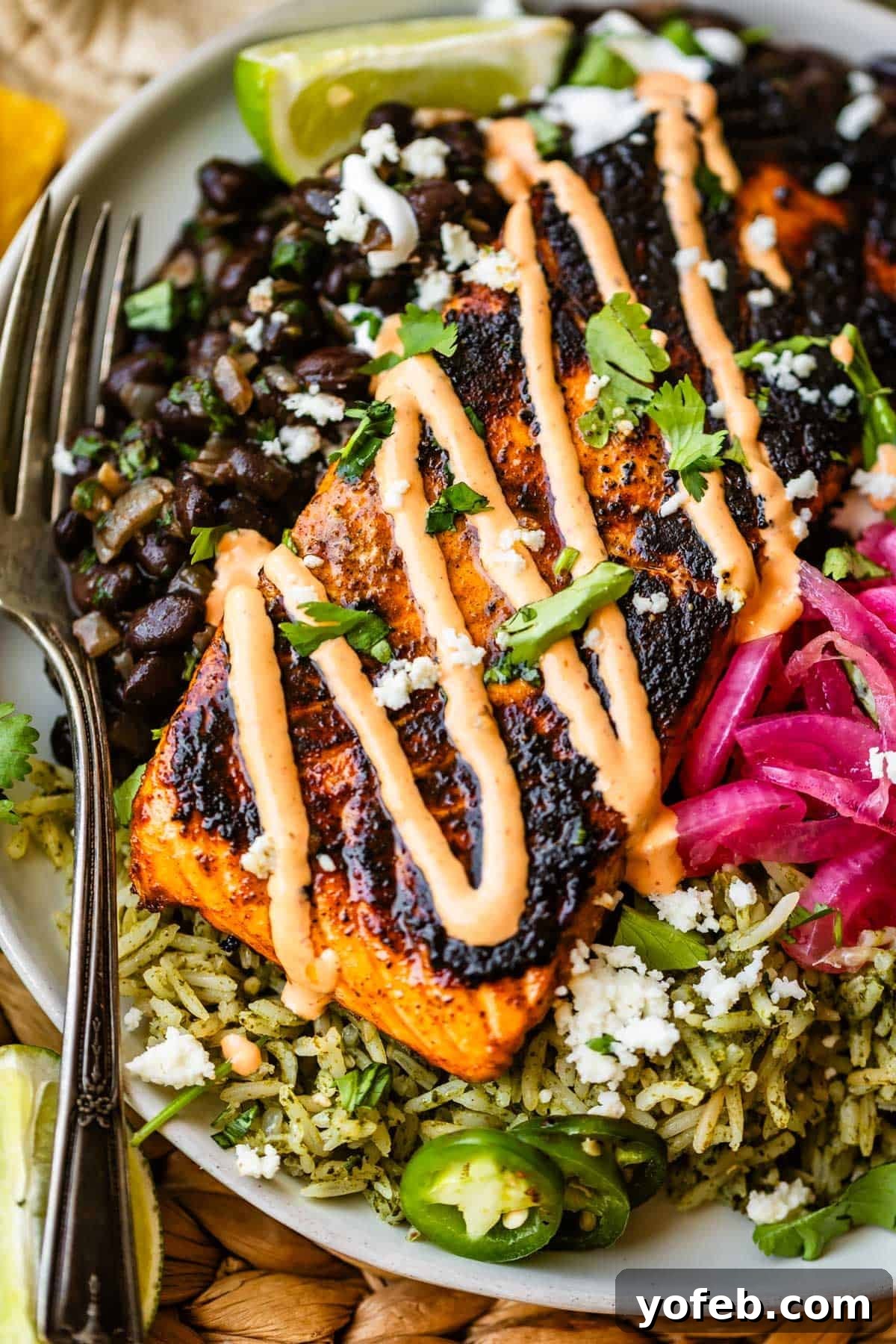 Chili-Lime Blackened Salmon 5 A grilled salmon fillet with sauce sits on green rice, next to black beans, pickled onions, jalapeños, cilantro, and lime wedges on a plate, with a fork on the side.