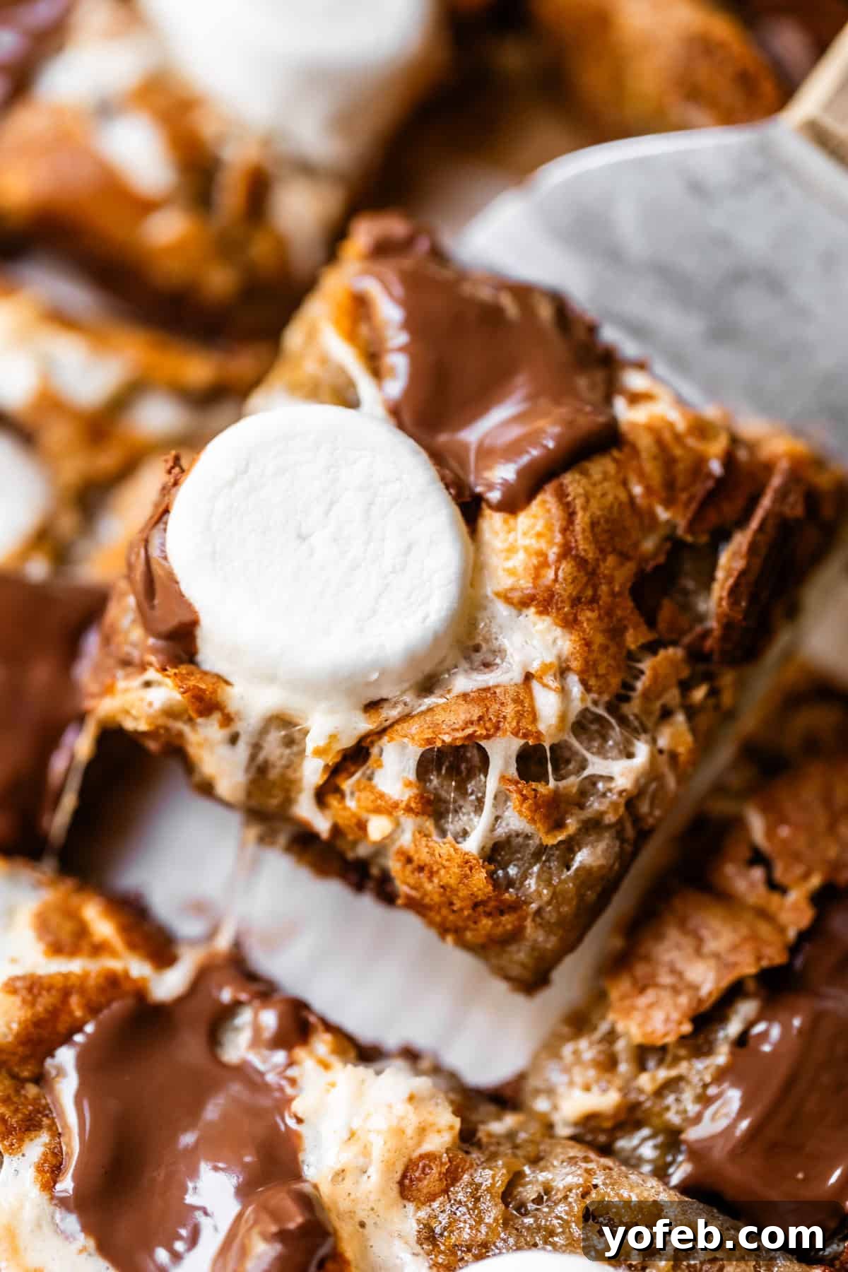 A close-up of a gooey smores bar topped with melted chocolate and a toasted marshmallow, being lifted by a spatula.