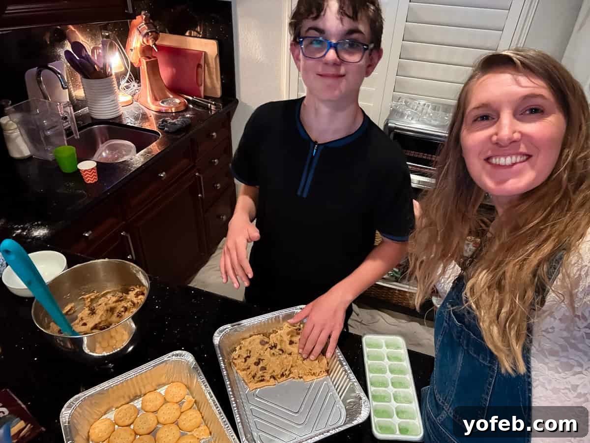 A woman and a boy smile while preparing cookie dough and graham crackers in a kitchen, with baking trays, bowls, and utensils on the counter.