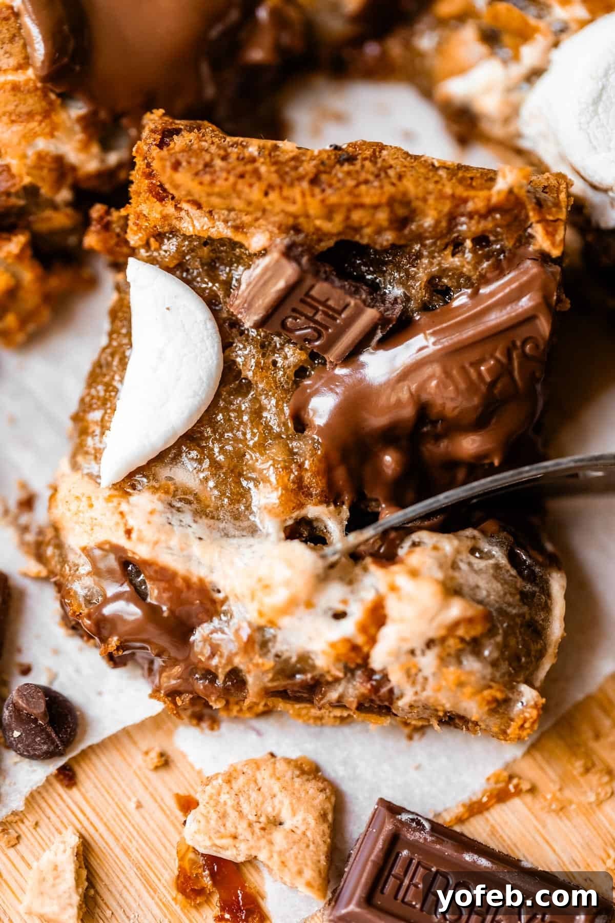 Close-up of a gooey smores bar topped with melted chocolate, graham cracker pieces, and toasted marshmallow, sitting on parchment paper with bits of chocolate and crumbs around it.