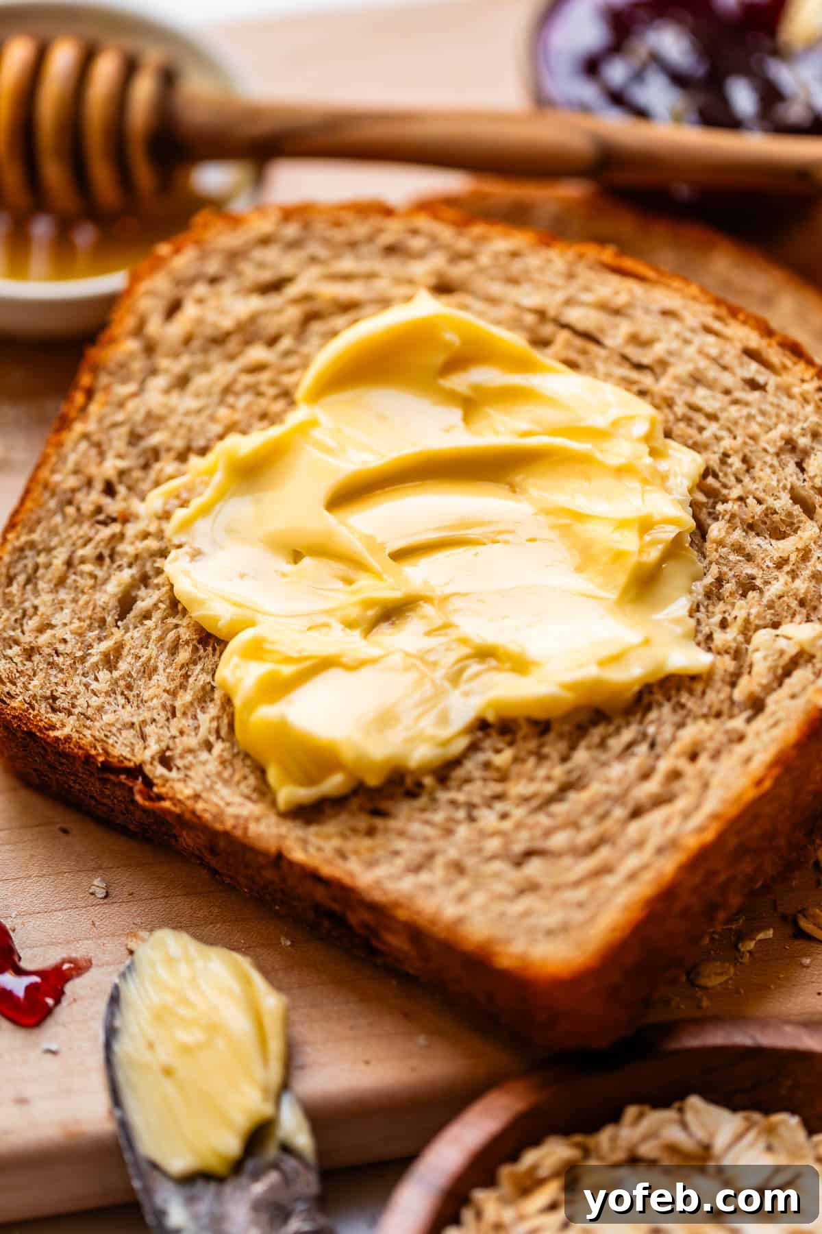 Soft Honey Whole Wheat Bread Recipe 2 A slice of whole grain bread with creamy butter spread on top, surrounded by a honey dipper, jam, and a butter knife with more butter.