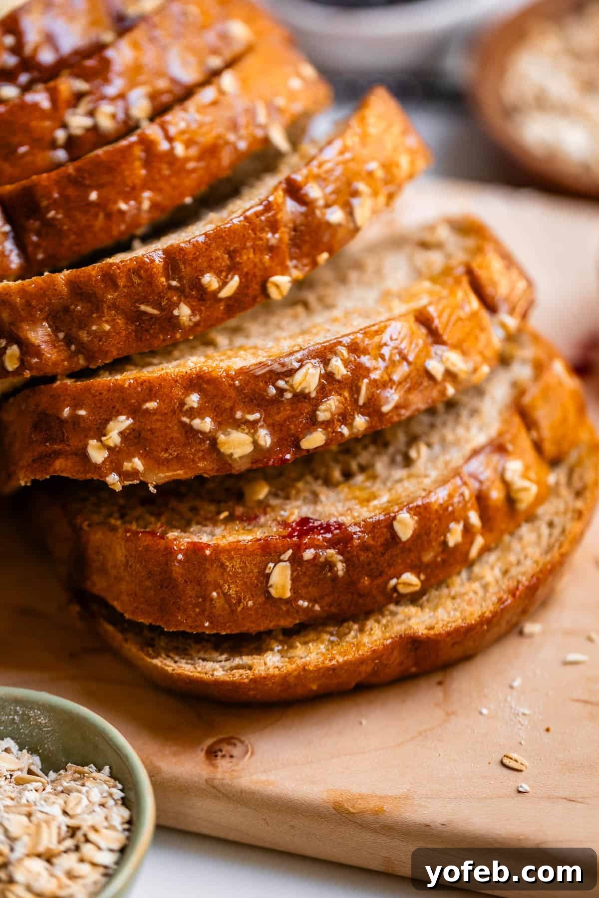 Soft Honey Whole Wheat Bread Recipe 3 Several slices of oat-topped bread are stacked on a wooden cutting board, with a small bowl of oats beside them. The bread looks freshly baked and golden brown.