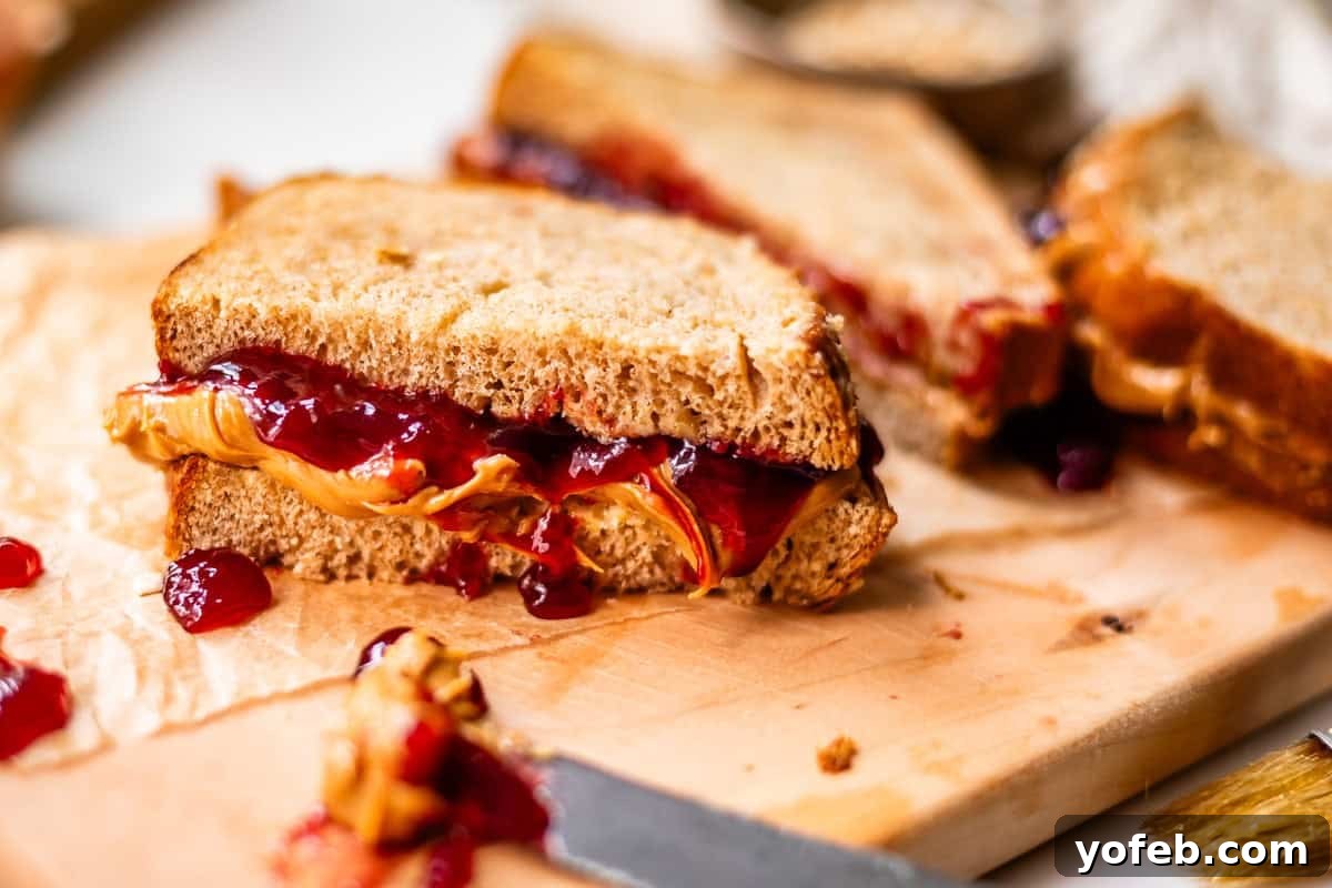 Soft Honey Whole Wheat Bread Recipe 4 A close-up of a peanut butter and jelly sandwich cut in half on a wooden cutting board, with peanut butter and jelly oozing from the sides.