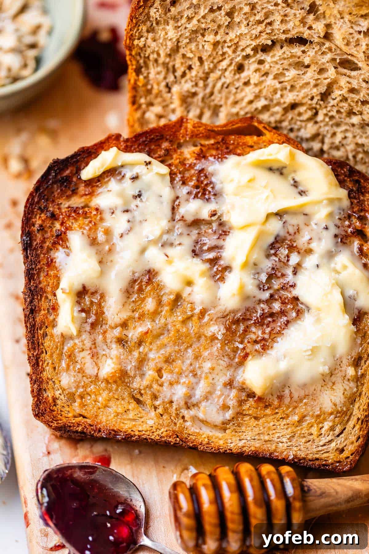 Soft Honey Whole Wheat Bread Recipe 15 A slice of toasted bread topped with melting butter, next to a loaf of bread, a honey dipper, and a spoon with dark fruit jam on a wooden board.