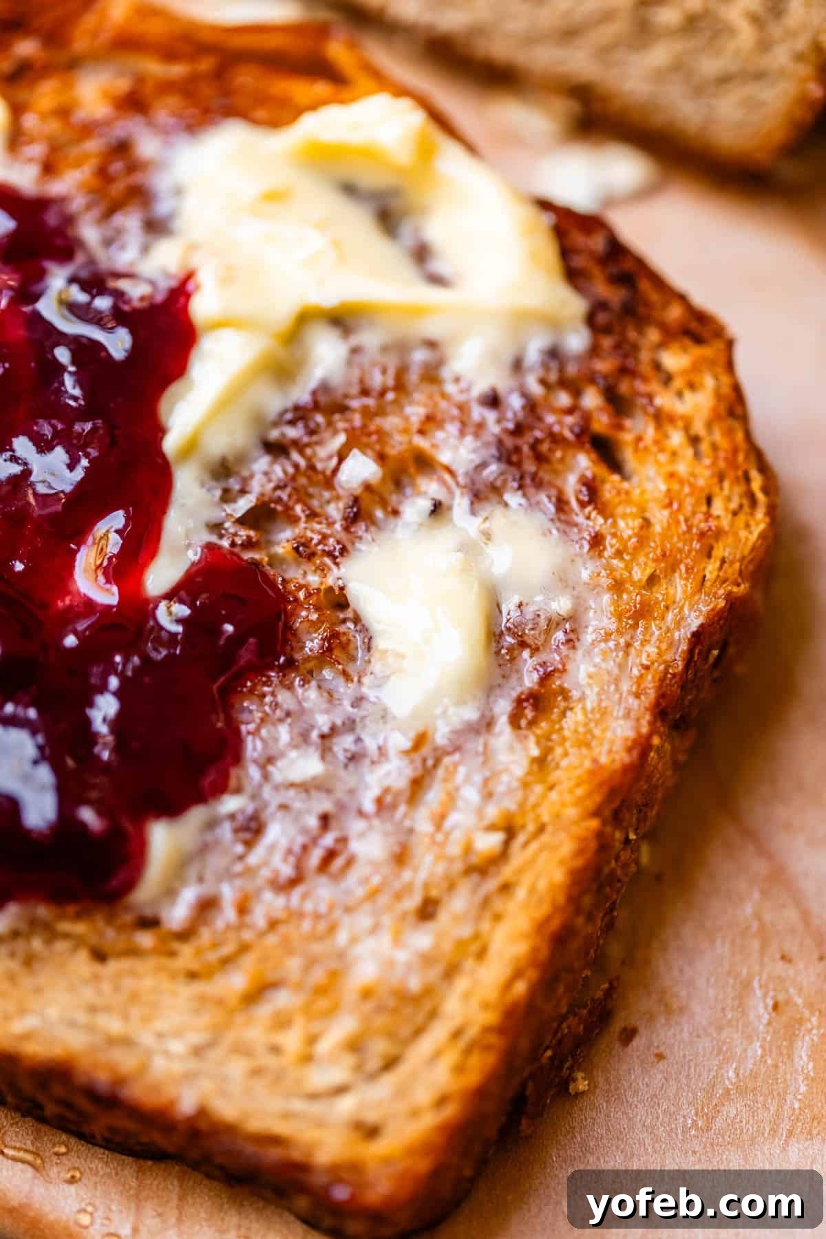 Soft Honey Whole Wheat Bread Recipe 6 A close-up of toasted bread spread with melting butter and red fruit jam, placed on a wooden surface.