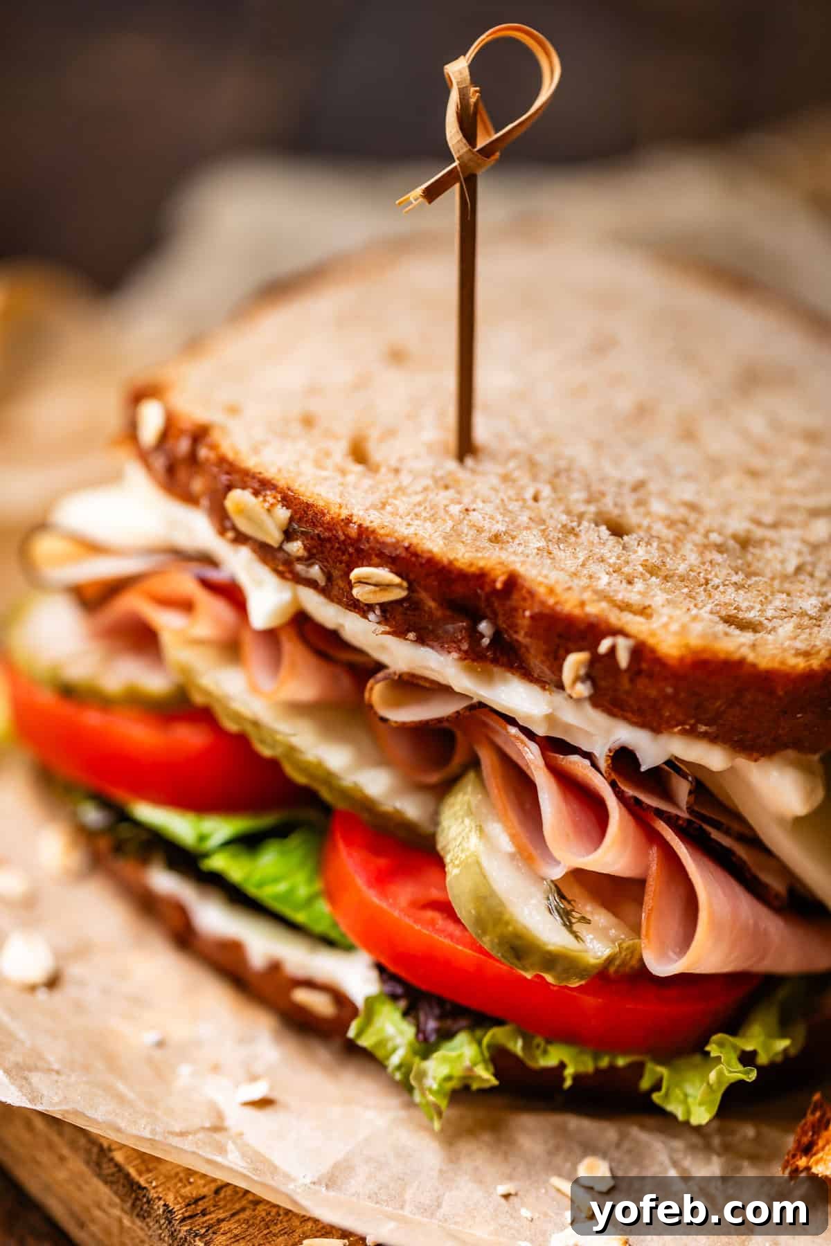 Soft Honey Whole Wheat Bread Recipe 7 Close-up of a sandwich on whole grain bread, filled with lettuce, tomato slices, pickles, ham, cheese, and mayonnaise, secured with a wooden pick.