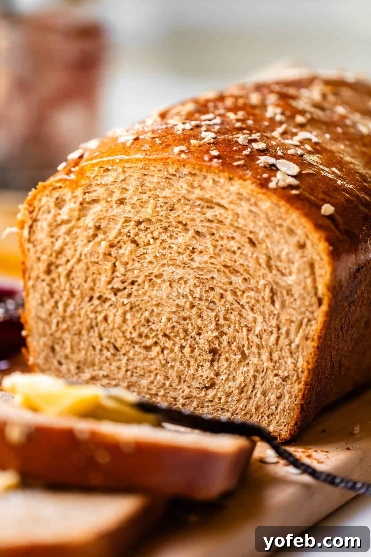 Soft Honey Whole Wheat Bread Recipe 8 A close-up of a loaf of whole wheat bread on a wooden board, with two slices cut and some butter and jam visible in the foreground. Oats are sprinkled on top of the loaf.