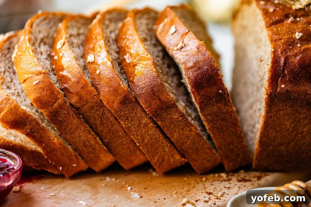 Soft Honey Whole Wheat Bread Recipe 10 A close-up of several slices of freshly baked bread arranged in a row on a wooden surface, with a loaf in the background and a small dish of jam partially visible beside them.
